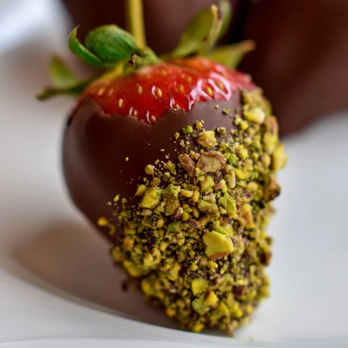 Fresh ripe strawberries dipped in glossy dark chocolate, sprinkled with crunchy chopped pistachios on a white plate.