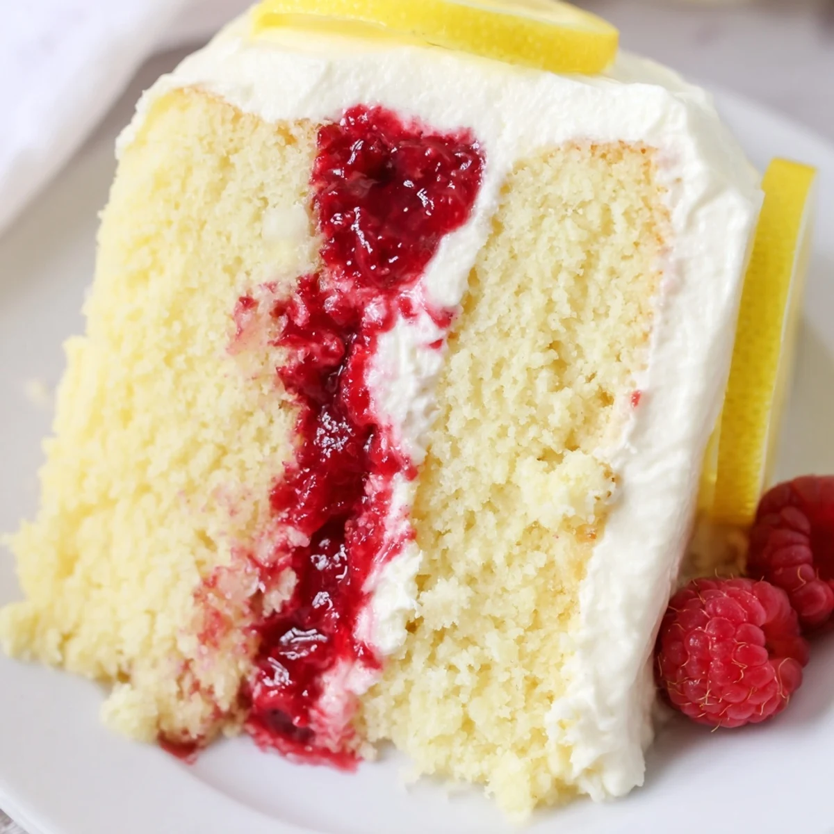 Moist Lemon Raspberry Layer Cake slices reveal tangy raspberry jam and lemon zest filling, topped with creamy frosting and fresh raspberries on a white plate.