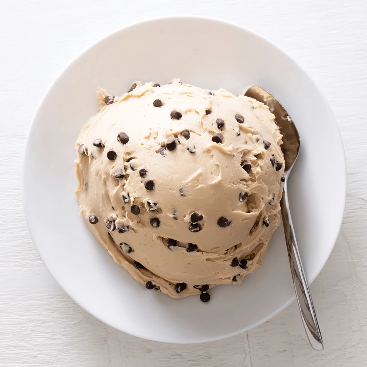 A bowl of freshly made Cookie Dough Frosting beside cupcakes, with a spoon ready for a sweet taste.