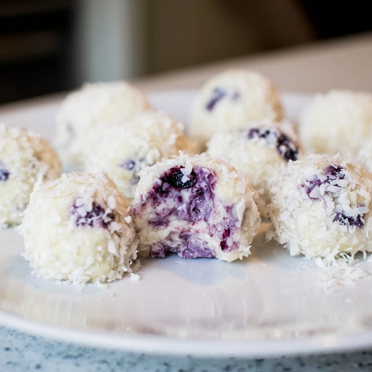 Creamy blueberry cheesecake protein bites rolled in coconut on a white serving plate.