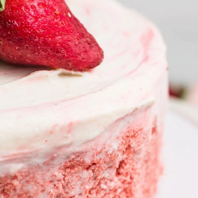 A whole Strawberry Velvet Cake on a marble stand, frosted with strawberry cream cheese frosting and topped with halved fresh strawberries for a spring celebration.
